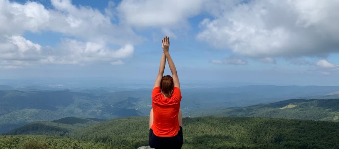 A woman in a red shirt relaxes on a mountain peak with arms raised, enjoying the scenic view.