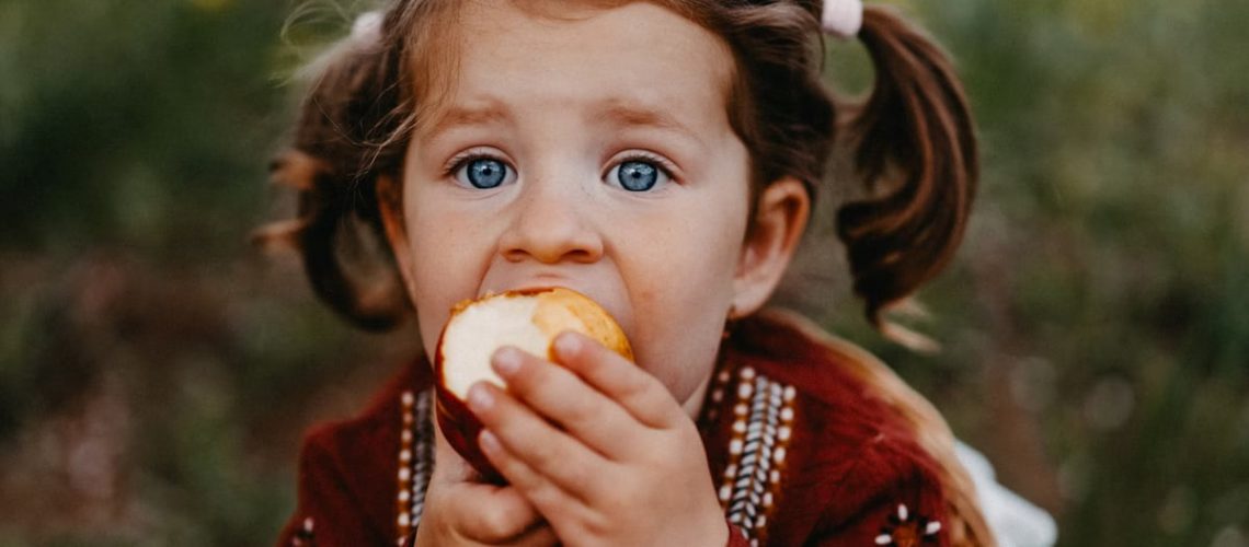 A young girl with ponytails enjoying an apple in a nature setting, captured in warm fall tones.